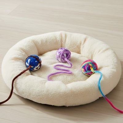 Balle en laine pour chat en trois coloris, bleu, violet et multicolore, sur un coussin beige